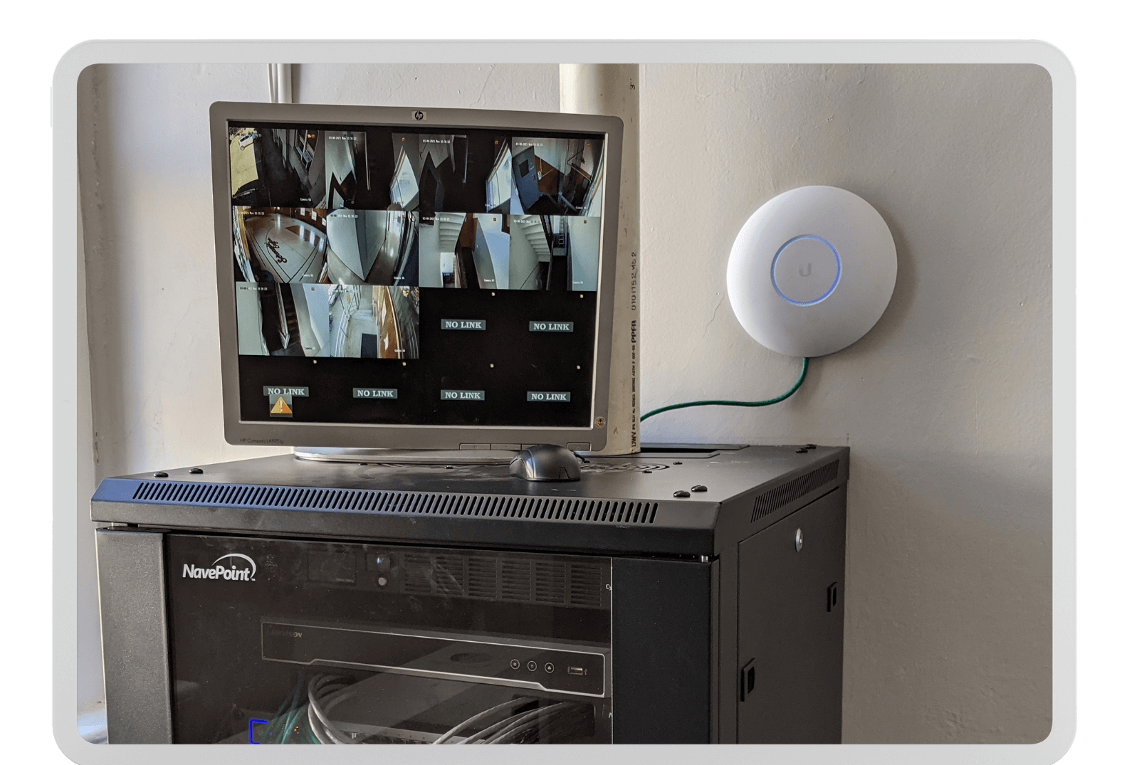 A small server rack with a monitor displaying multiple security camera feeds is mounted on top. The monitor shows various views of an indoor environment labeled "DINING LOUNGE." Next to the server rack, a round, white Ubiquiti networking device is mounted on the wall.