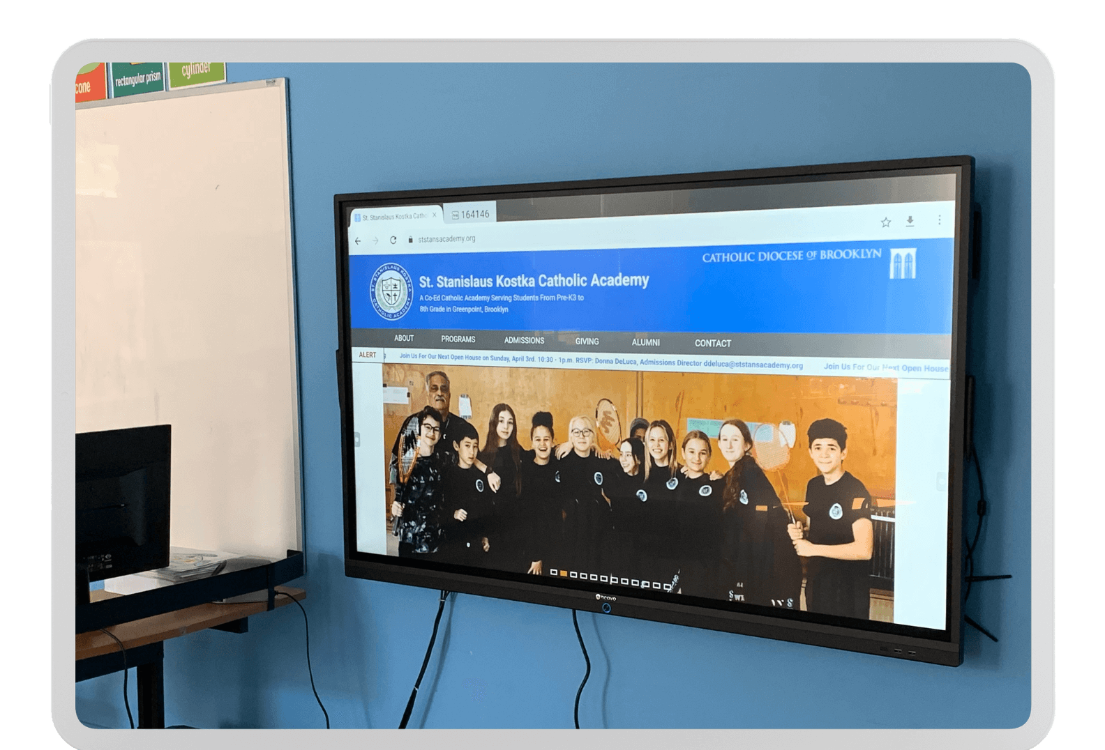 A large AG Neovo Interactive Flat Panel Display mounted on a blue wall showcases the homepage of the St. Stanislaus Kostka Catholic Academy website, featuring a group of people in black clothing posing together. A desk with a computer and keyboard is partially visible on the left.