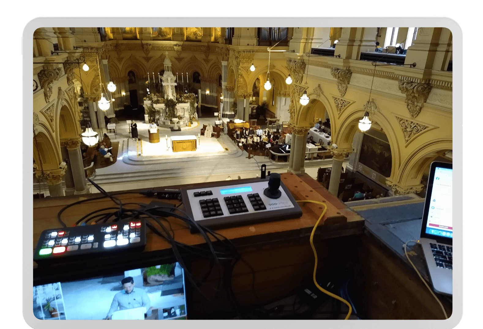 View from a balcony overlooking the interior of a church during a service. Various video equipment is set up on a table in the foreground, including a laptop, control panels, and screens. The ornate architecture and congregation are visible below.